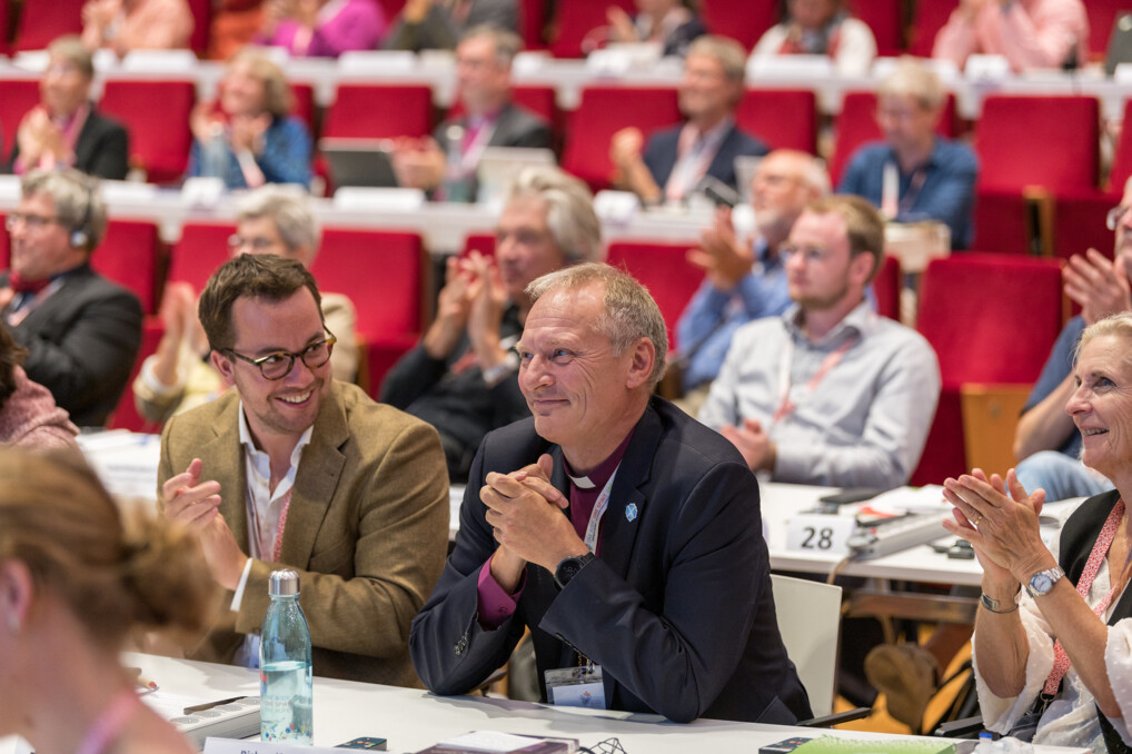 Billedet viser en gruppe mennesker, der sidder i et konferencelokale i Krakow i Polen med røde stole. Den kommende præsident Henrik Stubkjær sidder i midten af billedet. Nogle personer klapper, mens andre har hænderne sammen, muligvis i forventning eller applaus. Der er en flaske vand og papirer på bordene foran nogle af deltagerne. Rummet er godt oplyst og virker rummeligt.
