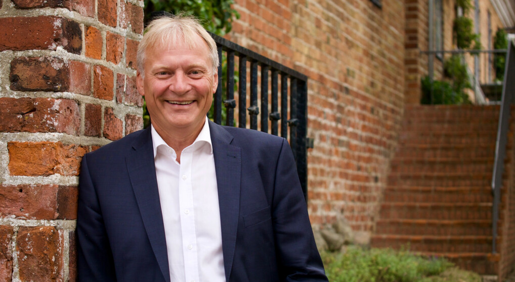 Bishop Henrik Stubkjær, dressed in a suit, stands next to a wall of red bricks. His face is blurred to protect privacy. The background reveals an old, weathered wall and a black metal railing along a staircase leading upward. 