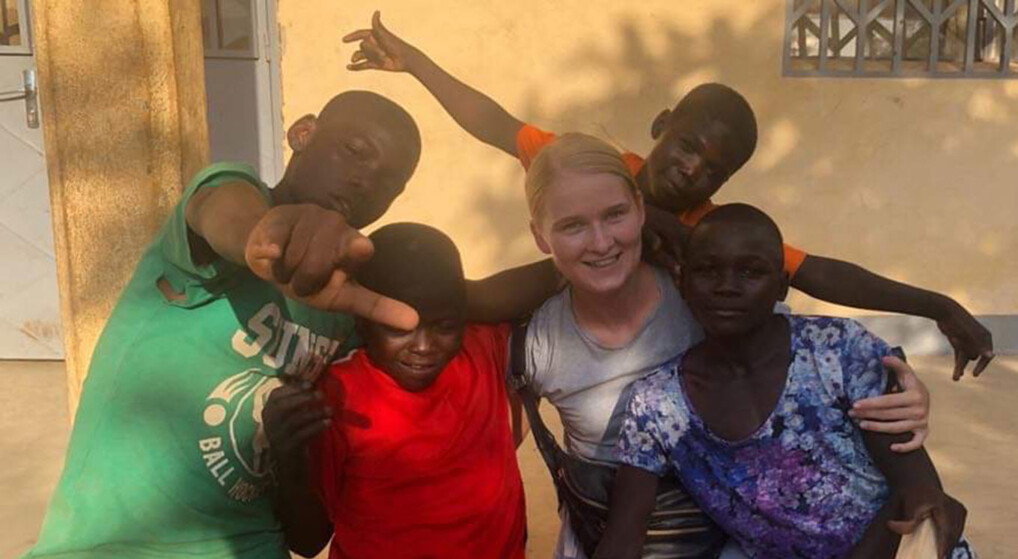 Billedet viser en gruppe på fem personer, der poserer sammen. De står udendørs, og baggrunden viser en mur og et vindue. En person har en grøn t-shirt med tekst og grafik, en anden er iført rødt tøj, en tredje bærer hvidt, og en fjerde har en mønstret blå beklædning. Deres arme er omkring hinanden, hvilket indikerer kammeratskab eller venskab. 